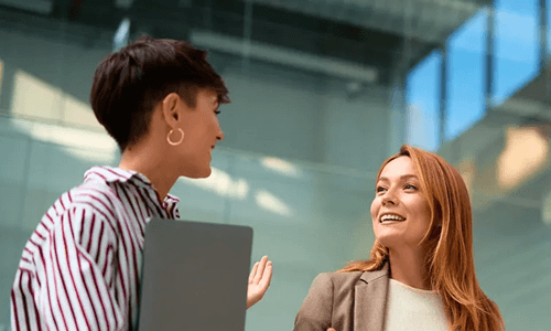 Dos colegas conversan animadamente de pie en un pasillo de oficina moderno, una con laptop y otra con tablet.