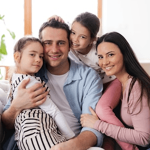 Familia de cuatro sonriendo abrazada en el sofá de su hogar: padre, madre y dos hijas pequeñas.